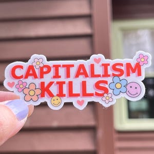 May include: A white sticker with the words "CAPITALISM KILLS" in red, decorated with flowers, hearts, and smiley faces. The sticker is held by a hand with a light purple painted nail.