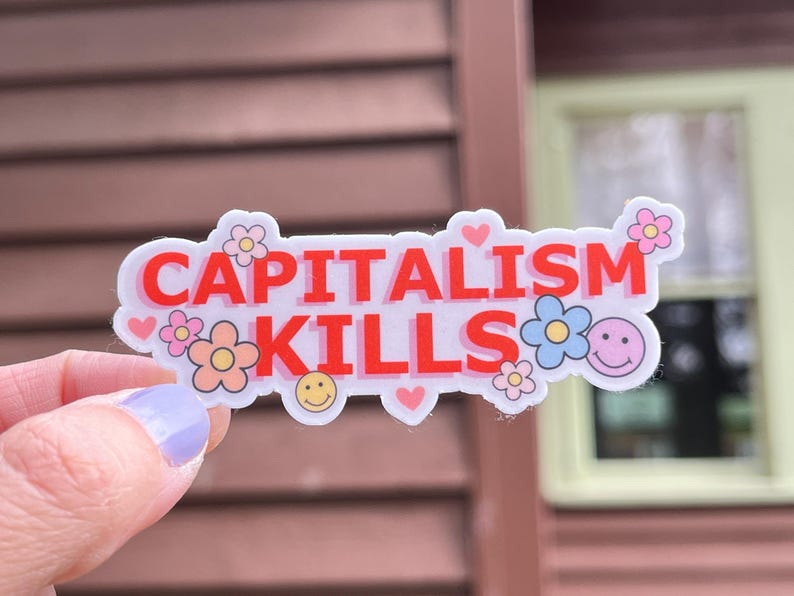 May include: A white sticker with the words "CAPITALISM KILLS" in red, decorated with flowers, hearts, and smiley faces. The sticker is held by a hand with a light purple painted nail.