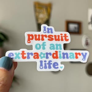 Puede incluir: Una pegatina blanca con el texto "In pursuit of an extraordinary life" en naranja, azul y morado. La pegatina tiene una forma única y está sujeta por una mano con una uña pintada de azul.