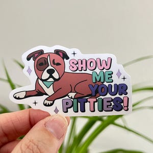 May include: A sticker featuring a cartoon brown and white pit bull dog with a pink, green, and purple collar. The text "SHOW ME YOUR PITTIES!" is written in pink, green, and purple letters.