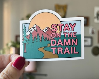 May include: A rectangular sticker with a landscape design. The sticker features a mountain range, trees, a winding path, and a setting sun. The text "STAY ON THE DAMN TRAIL" is in pink.