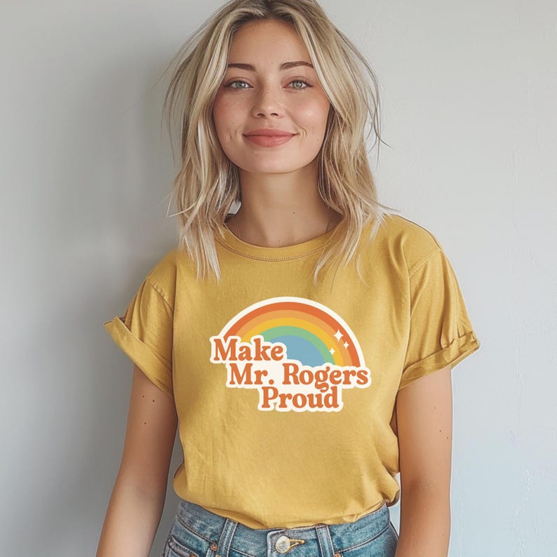 May include: Mustard yellow t-shirt with rolled-up sleeves. The shirt features a rainbow graphic and the text "Make Mr. Rogers Proud" in a retro font. The shirt is made of a soft material.