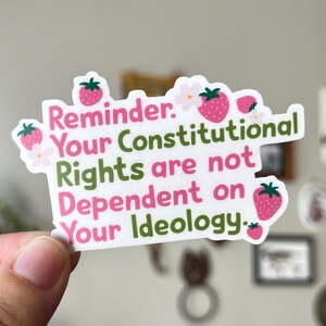 May include: A white sticker with a strawberry and flower design. The sticker has the text "Reminder: Your Constitutional Rights are not Dependent on Your Ideology" in pink and green.