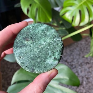 Puede incluir: Una pastilla de jabón redonda, verde y blanca con textura moteada. El jabón está en la mano de una persona.