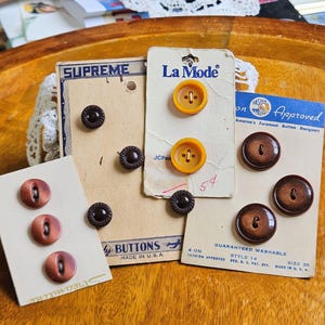May include: A collection of vintage buttons on their original cards. The buttons vary in color, including brown, amber, and reddish-brown. Some cards are labeled "SUPREME", "La Mode", and "Fashion Approved". The image showcases a variety of sizes and styles.