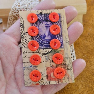 May include: A collection of ten bright orange, round buttons with two holes, attached to a vintage postcard. The buttons are a vibrant, textured orange. The postcard has a faded, antique design with stamps and script.