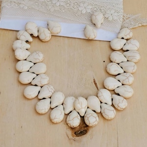 May include: A white teardrop-shaped beaded necklace with a delicate, marbled pattern. The necklace is made of natural stone beads and is strung on a white cord.