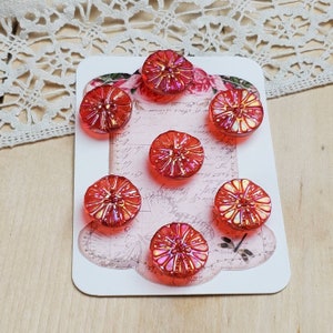 May include: Seven red glass buttons with a flower-like design are arranged on a decorative card. The buttons have a textured surface and iridescent highlights. The card has a floral and script design.