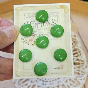 May include: Six round, glossy, grass-green buttons arranged on a rectangular display card. The card features a vintage-style floral design with the text "Buttons" and "Size 15" printed in black ink. Each button has a single small hole for attachment.