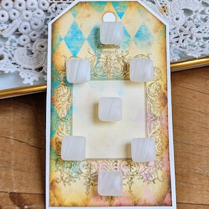 May include: A collection of six square, white, marbled glass buttons arranged on a decorative tag. The tag has a vintage design with a diamond pattern in blue and gold, and ornate scrollwork. The buttons are approximately 0.5 inches square.