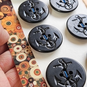 May include: A collection of black buttons with an anchor design and blue accents. The buttons are round and have four holes for sewing. A decorative strip with a colorful floral pattern is also visible.
