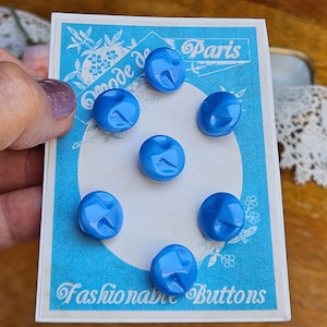 May include: Six round, bright blue buttons with a smooth, glossy finish and a subtle indented detail, arranged on a rectangular light blue display card featuring the text Mode de Paris and Fashionable Buttons in white script and print.