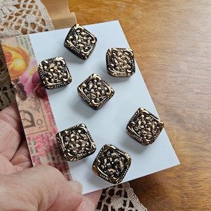 May include: Seven square, antique-style buttons with a gold-colored border and a textured, gold-colored center. The buttons are arranged on a white card, with a floral design and text visible in the background.