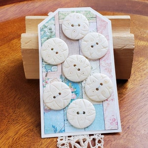 May include: Cream-colored, round buttons with a floral design are arranged on a decorative tag. The tag features a collage of pastel floral and patterned papers. A wooden block and lace trim are also visible.