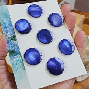 May include: Seven round, iridescent blue buttons with a swirling pattern are arranged on a white card. The buttons have a glossy finish and vary slightly in the intensity of their blue hues. The card is held by a person's hand, with a floral design visible on the left side.
