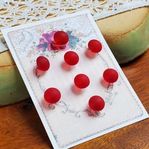 May include: A collection of eight small, round, red glass beads arranged on a decorative card. The card features a floral design with a vintage aesthetic. The beads are evenly spaced, creating a visually appealing pattern. The background includes lace and a green object.