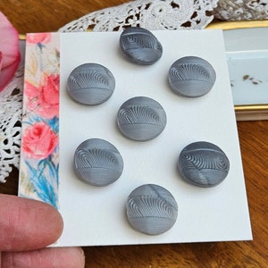 May include: A collection of seven gray buttons with a textured, wave-like design. The buttons are round and appear to be made of a hard material. They are arranged on a white card, with a floral patterned edge visible.