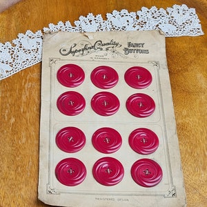 May include: A vintage card of twelve red, spiral-design buttons. The card reads "Superfine Quality Fancy Buttons Made in Germany." A white lace trim is in the upper left corner. The buttons are a deep red color and have a small hole in the center.