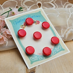 May include: A card of vintage red buttons. The buttons are round and glossy, arranged in a circular pattern on a card with the words "Vintage Buttons" and floral accents. The card has a light blue border and is on a wooden surface.
