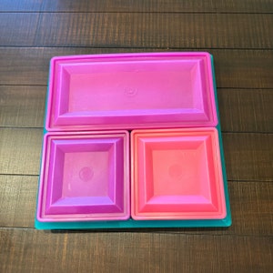 Puede incluir: Un juego de cuatro recipientes de almacenamiento de plástico con tapas. Los recipientes están dispuestos en forma de cuadrado. El recipiente superior es morado, los dos recipientes inferiores son morado y rosa, y la base es turquesa.