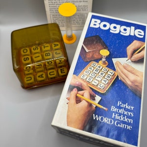 May include: A close-up of the Boggle board game. The box is blue with white lettering and shows people playing the game. The game includes a letter cube tray, a timer, and a score sheet. The game is called "Parker Brothers Hidden Word Game".