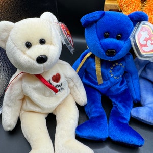 May include: Two plush teddy bears, one white with a red "I Love New York" heart on its chest and the other blue with gold stars and a yellow ribbon around its neck.