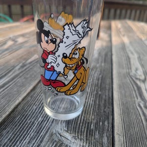 May include: A clear glass featuring Mickey Mouse and Pluto cartoon graphics. Mickey is wearing a red shirt, blue pants, and white gloves. Pluto is a yellow dog with a red collar. The glass is cylindrical and sits on a wooden surface.