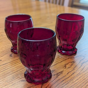 May include: Three ruby red glass tumblers with a textured, faceted design. The glasses are sitting on a wooden table.