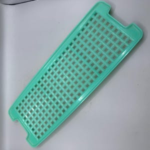 May include: A rectangular, light green plastic kitchen utensil with a grid pattern of square holes. The edges are slightly raised, and it has two small handles on one end. The item is designed for draining or rinsing food items.