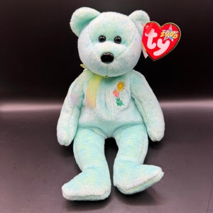 May include: A light blue Ty Beanie Baby bear named "Spring" with a yellow ribbon around its neck and embroidered flowers on its chest. The bear is sitting with its feet tucked under its body. The bear has a red Ty heart tag with the year 2000 printed on it.