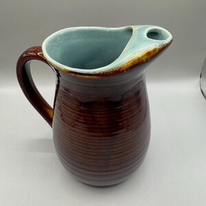 May include: A brown ceramic pitcher with a handle and a unique spout design. The interior is a light blue color, and the rim has a brown and gold trim. The exterior has a textured, ribbed pattern.