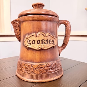 May include: Brown ceramic cookie jar with a lid and handle. The jar features the word "Cookies" in a decorative plaque. Floral and leaf patterns are carved around the base. The jar is on a wooden surface.