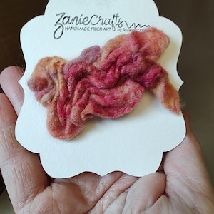 May include: A textured, felted fiber art brooch in shades of pink, orange, and purple, attached to a white scalloped display card with the text ZanieCrafts HANDMADE FIBER ART by Suzanne O'Pry printed in black ink at the top.