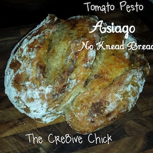 May include: A loaf of Asiago no-knead bread with tomato pesto, dusted with flour, on a wooden cutting board. The text on the image reads "Tomato Pesto Asiago No Knead Bread" and "The Creative Chick".
