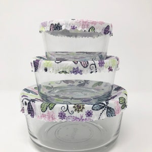 May include: Three glass bowls with floral patterned fabric covers. The covers have a white background with purple, green, and pink flowers and butterflies. The bowls are stacked on top of each other.
