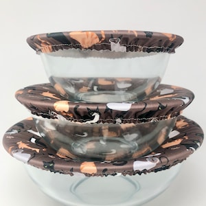 以下が含まれることがあります： 猫の繰り返し模様が施された、茶色と白の布製のカバーが付いたガラス製のボウルが3つ。カバーは柔らかく伸縮性のある素材でできており、フリルが付いています。