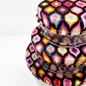 May include: Three reusable silicone food covers in a stack. The covers are made of a stretchy fabric with a geometric pattern in shades of pink, purple, orange, and yellow. The covers are designed to fit over bowls and containers to keep food fresh.