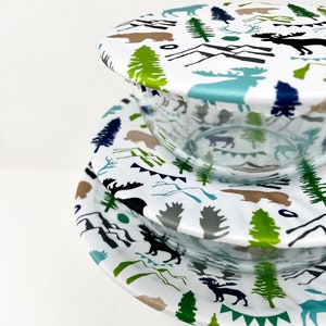 May include: Three reusable silicone food covers with a white background and a woodland pattern featuring trees, mountains, moose, and deer. The covers are in various sizes and are stacked on top of each other.