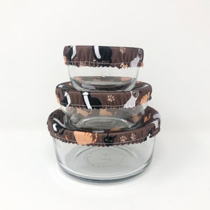 May include: Three glass bowls with brown fabric lids featuring a cat and paw print design. The lids are elastic and fit snugly over the bowls.