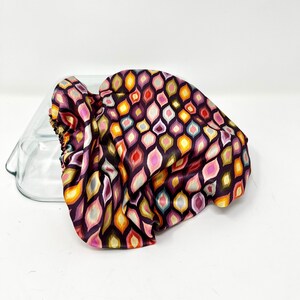 以下が含まれることがあります： カラフルな模様のついたベーキングディッシュ用の布製カバー。カバーは柔らかく絹のような素材でできており、濃い紫色の背景にピンク、オレンジ、イエロー、ブルー、グリーンの色の幾何学模様が施されています。