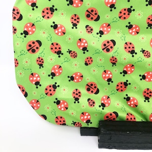 以下が含まれることがあります： 緑色の生地に、黒い斑点と白いアクセントのある赤いテントウムシが繰り返しパターンで描かれています。