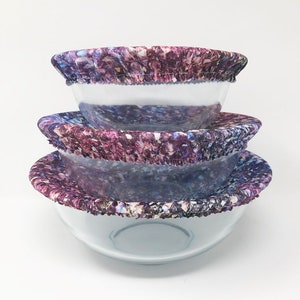 May include: Three reusable silicone bowl covers in a purple and blue abstract print. The covers are made of a stretchy material and can be used to cover bowls of different sizes.