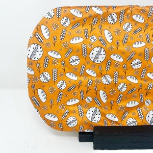 Könnte beinhalten: Ein orangefarbener Stoff mit einem sich wiederholenden Muster aus weißem Brot, Weizenähren und floralen Elementen. Der Stoff wird auf einem schwarzen Ständer präsentiert, was einen Kontrast zum hellen Design bildet. Das Thema bezieht sich auf Backen oder Essen.