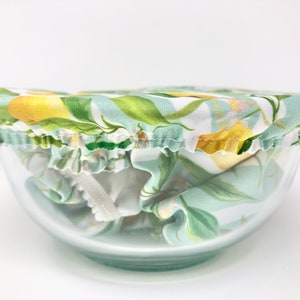 May include: A reusable bowl cover with a lemon and leaf pattern on a light blue background. The cover is made of fabric and has a ruffled edge. It is covering a clear glass bowl.