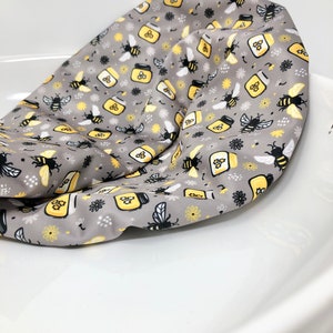 May include: A gray fabric cushion with a repeating pattern of black bees, yellow honey jars, and white flowers.