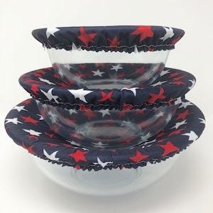 以下が含まれることがあります： 透明なガラスボウルが3つ重ねられており、それぞれに紺色の布製の蓋が付いています。蓋には赤と白の星模様が施されています。ボウルはサイズが異なり、一番大きいものが一番下にあります。