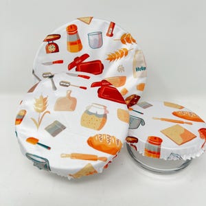 Peut inclure: Des couvercles de bols en tissu blanc avec un motif coloré sur le thème de la pâtisserie. Le motif comprend des images de pain, de rouleaux à pâtisserie, de balances de cuisine et de bouteilles de farine. Les couvercles sont conçus pour s'adapter aux bols et aux récipients.