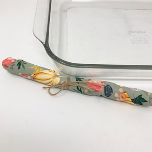 以下が含まれることがあります： 透明なガラス製のベーキングディッシュと、紐で結ばれた、丸められた模様入りの布アイテム。布には、カボチャ、葉、その他の秋の要素が描かれた花柄のデザインが施されています。