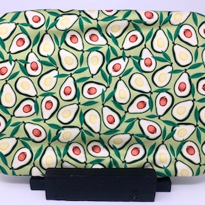 Puede incluir: Una bandeja de tela verde de forma ovalada con un patrón repetido de mitades de aguacate con huesos rojos y hojas verdes. La bandeja está sobre un soporte de madera negro.