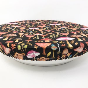 May include: A black bowl cover with a colorful mushroom pattern. The cover is stretched over a white bowl, showcasing a variety of mushroom illustrations in shades of red, orange, pink, and green. The cover is made of fabric.
